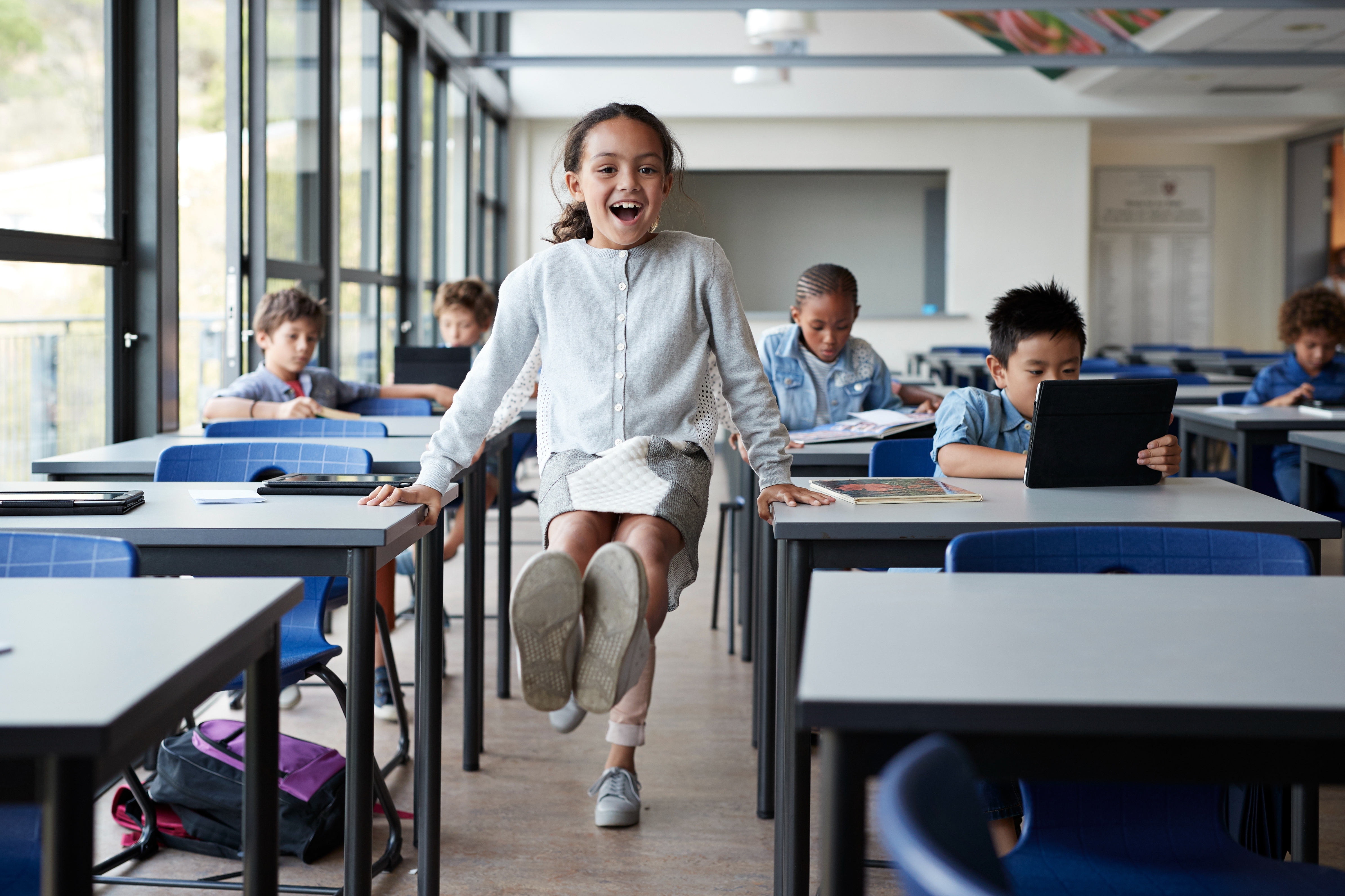 A child joyfully jumps in a classroom while other students work on tablets at their desks