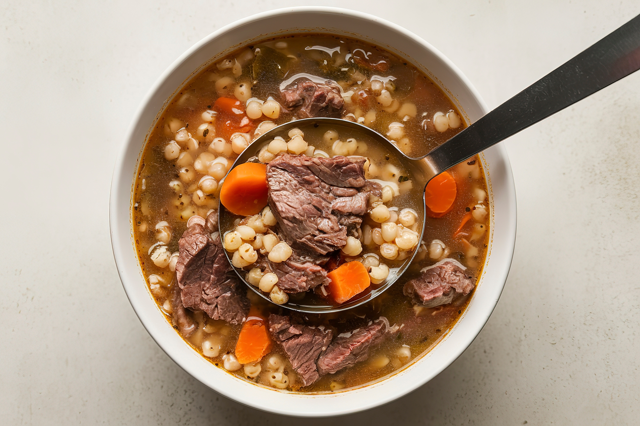 A bowl of beef and barley soup with chunks of beef, barley, carrots, and broth, shown with a ladle serving a portion