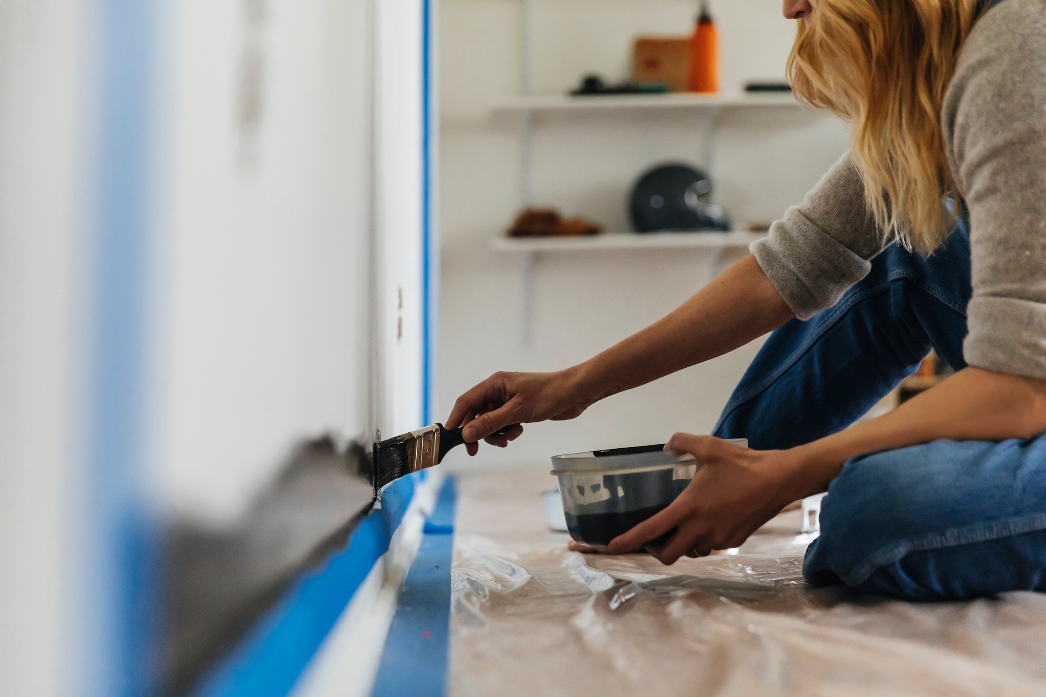 Person painting a wall with a brush, kneeling on a plastic-covered floor, holding a paint can. Shelves and items are visible in the background