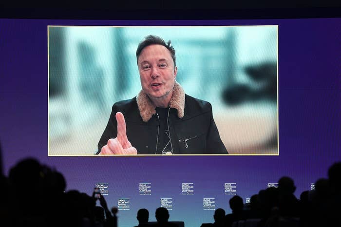 Elon Musk speaking on screen during a conference, wearing a jacket with a shearling collar. Audience silhouettes visible in the foreground