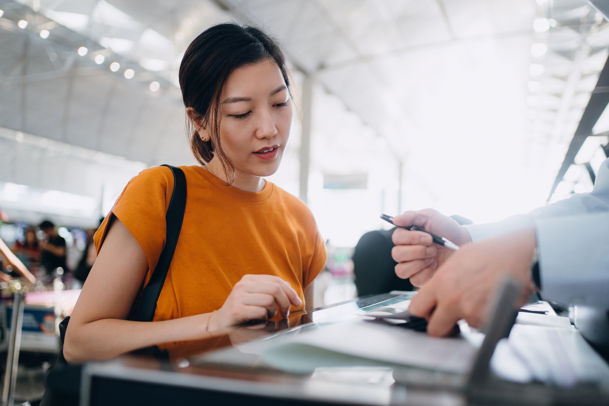 Woman at an airport check-in counter, watched by an employee who hands her a pen. She appears focused, with a casual style