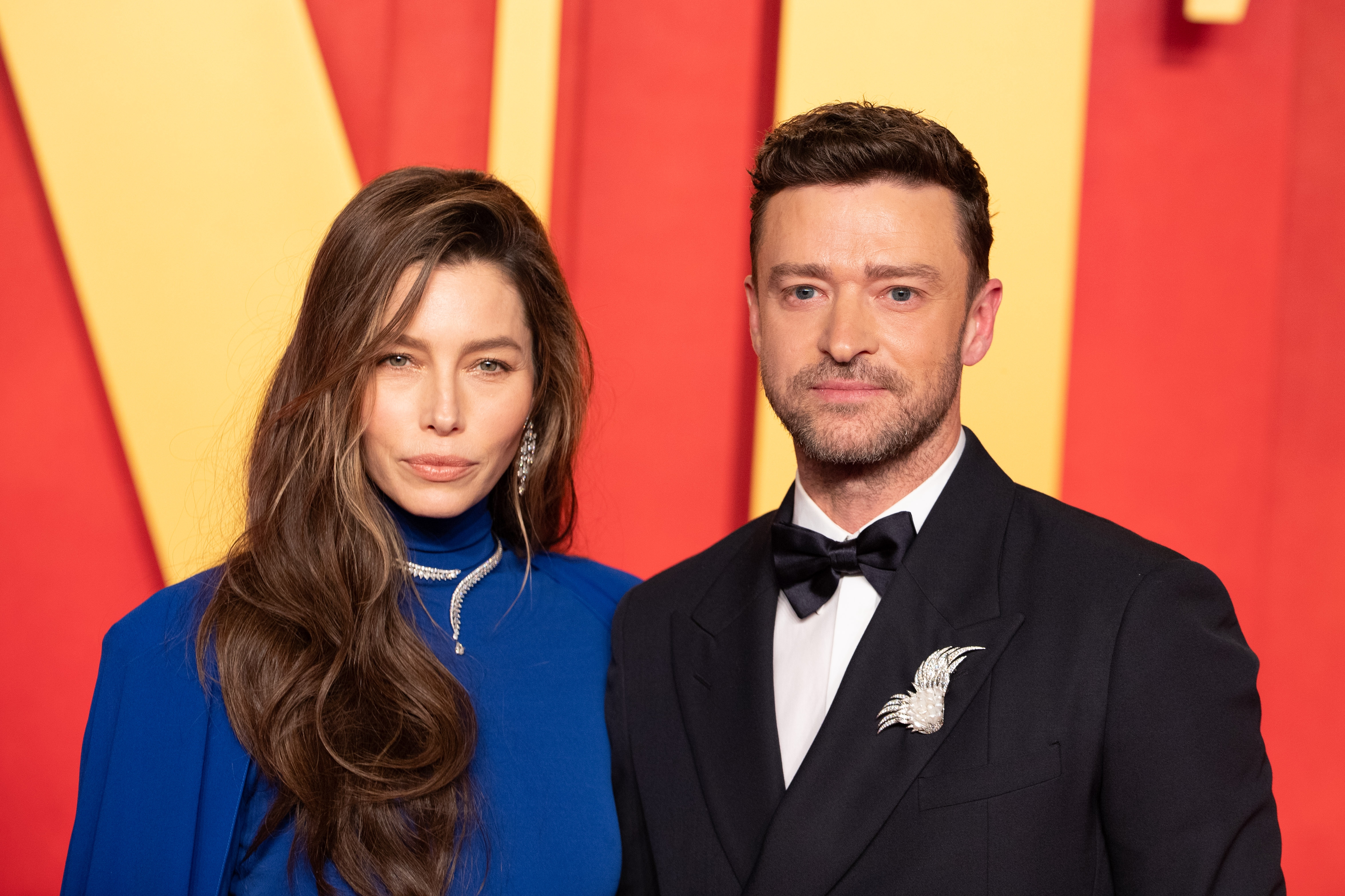 Jessica Biel and Justin Timberlake on a red carpet; one in an elegant blue dress with long hair, the other in a classic tuxedo with a bird-shaped brooch