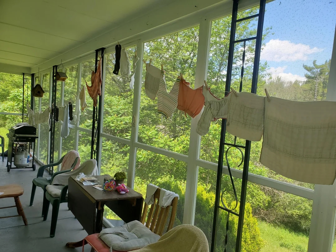 Clothes hanging on a line across a sunlit porch, with chairs and a table beneath them, overlooking a lush garden