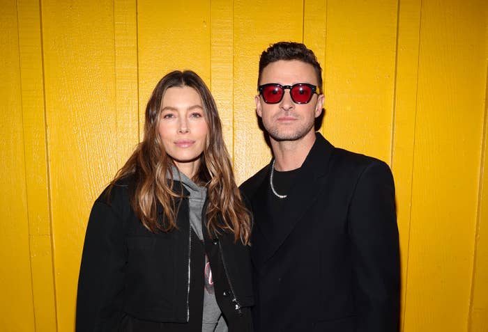 Jessica Biel and Justin Timberlake stand against a yellow wall. One wears a dark jacket; the other is in a black suit with red sunglasses