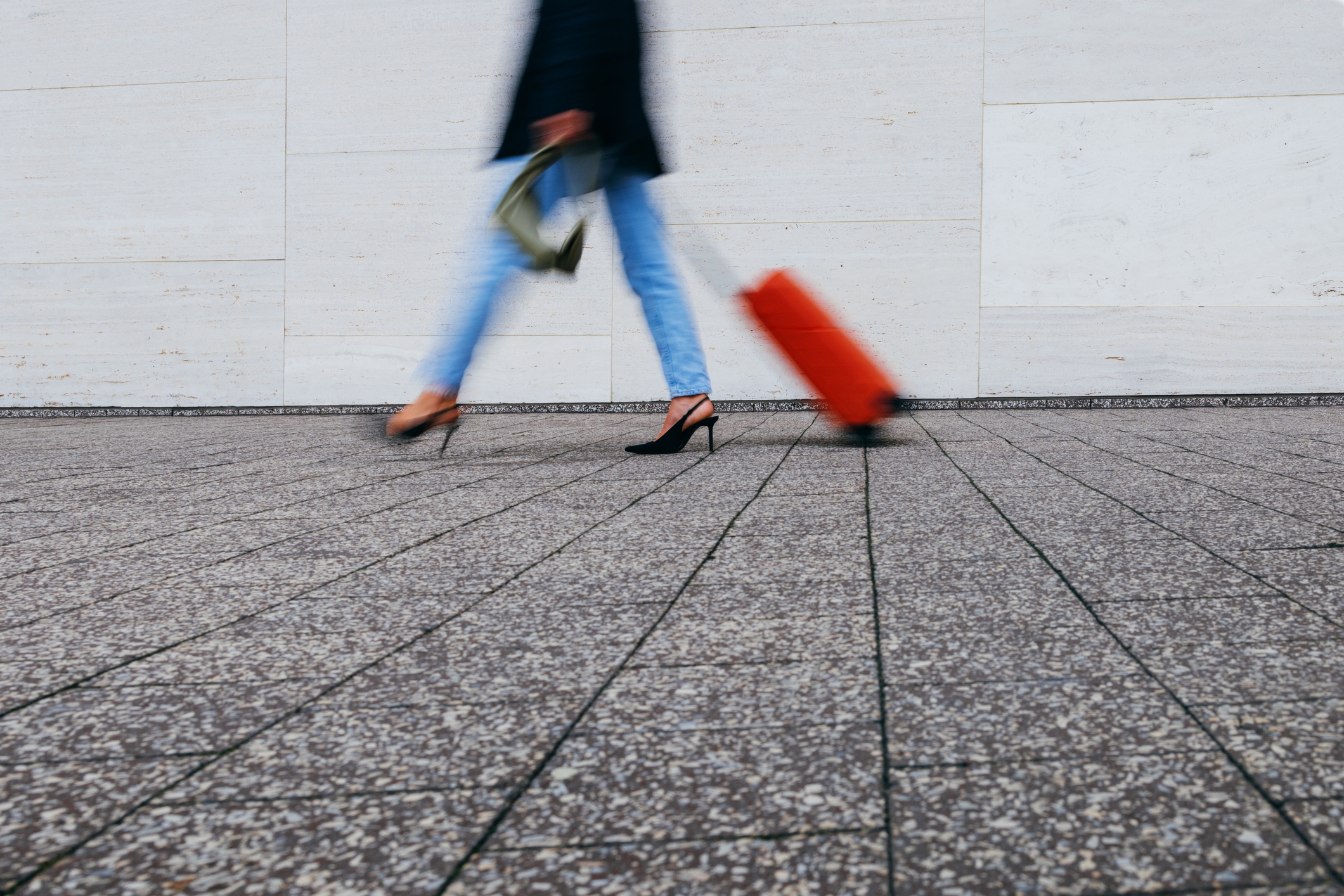 Person in heels walks briskly with a rolling suitcase, motion blur conveying speed, on a stone-paved area against a plain wall