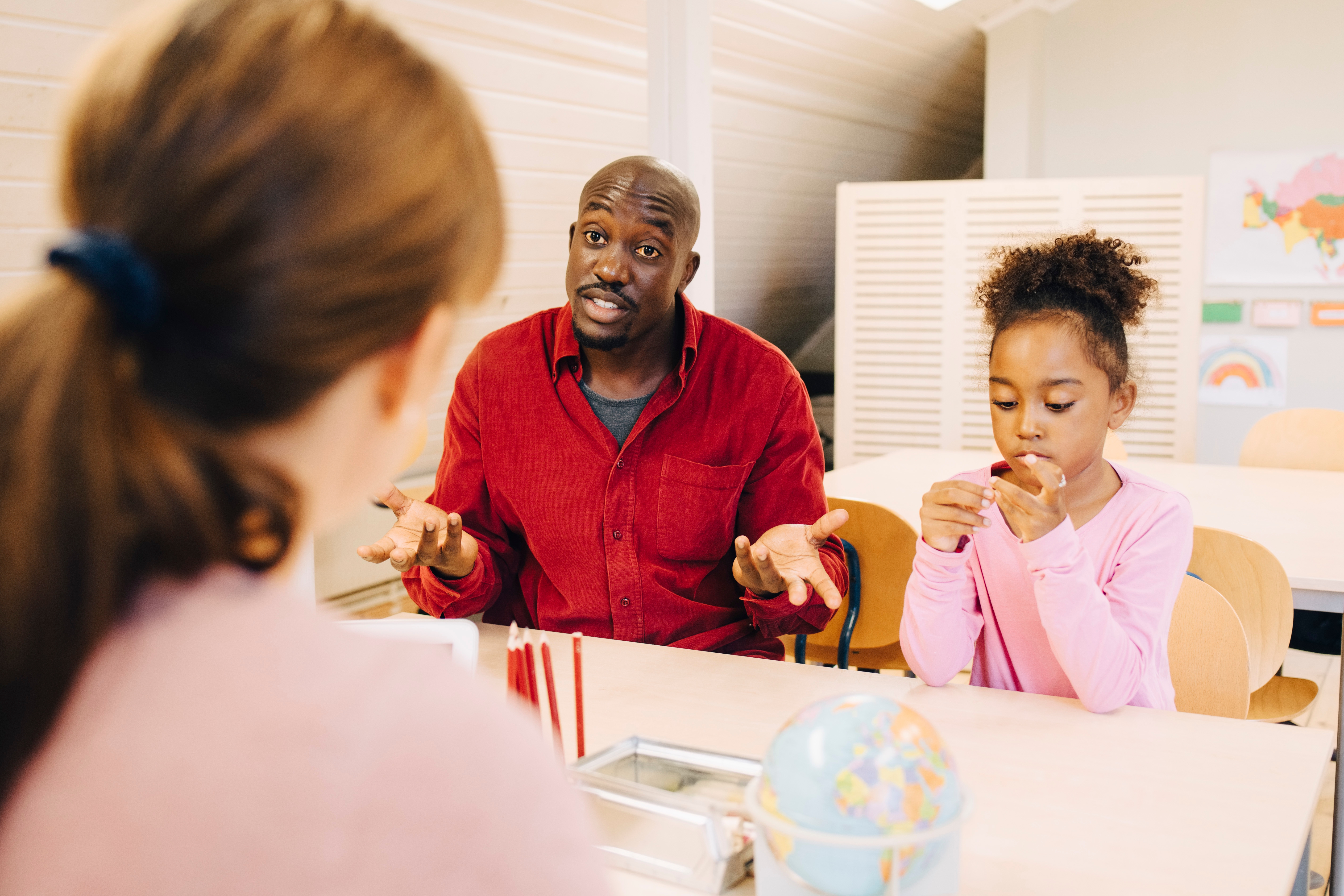 A man in casual attire talks to a woman at a table while a child focuses on a toy, suggesting a discussion about work-life balance
