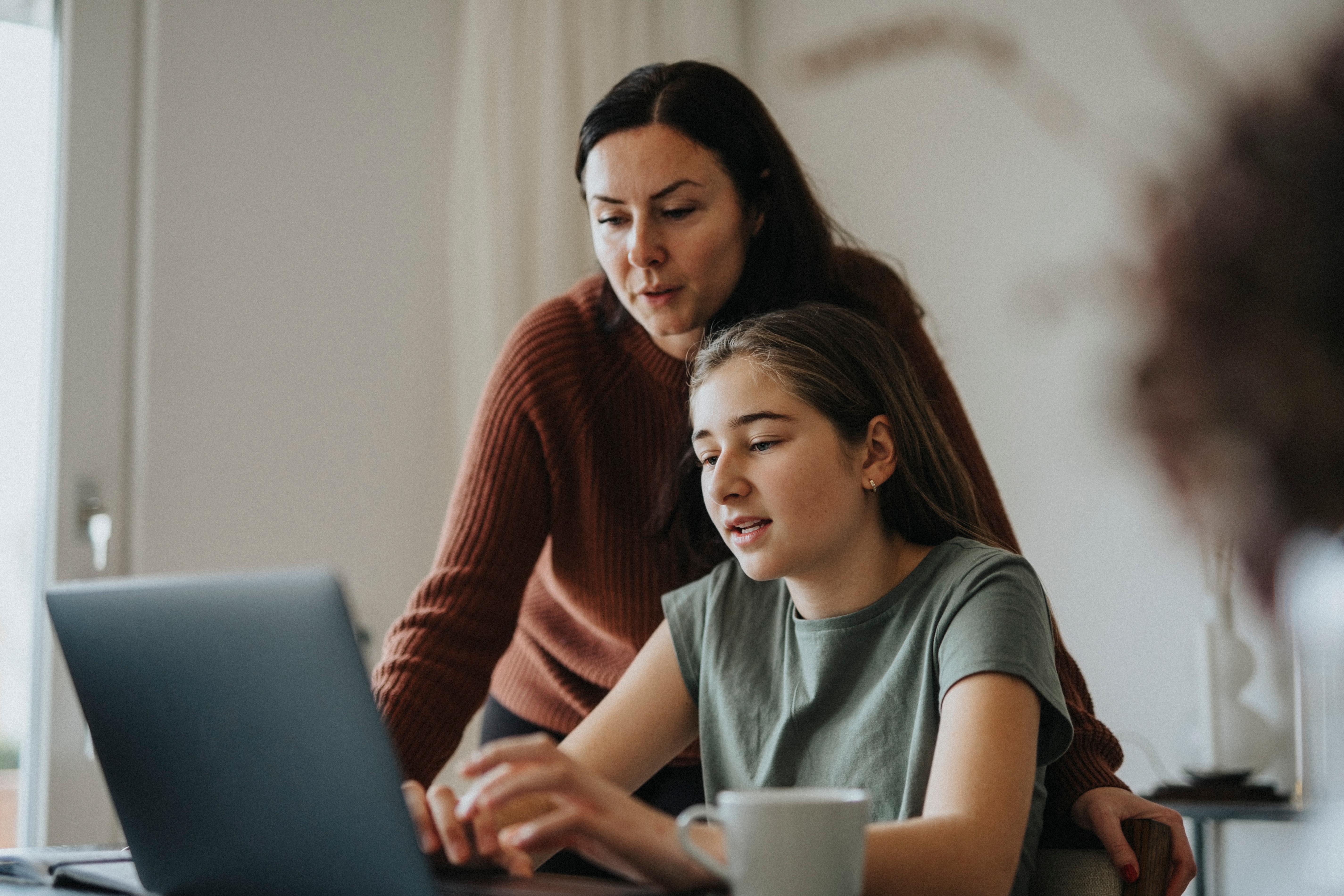 Teenager using a laptop, with an adult woman leaning over to assist, suggesting a work or learning environment at home