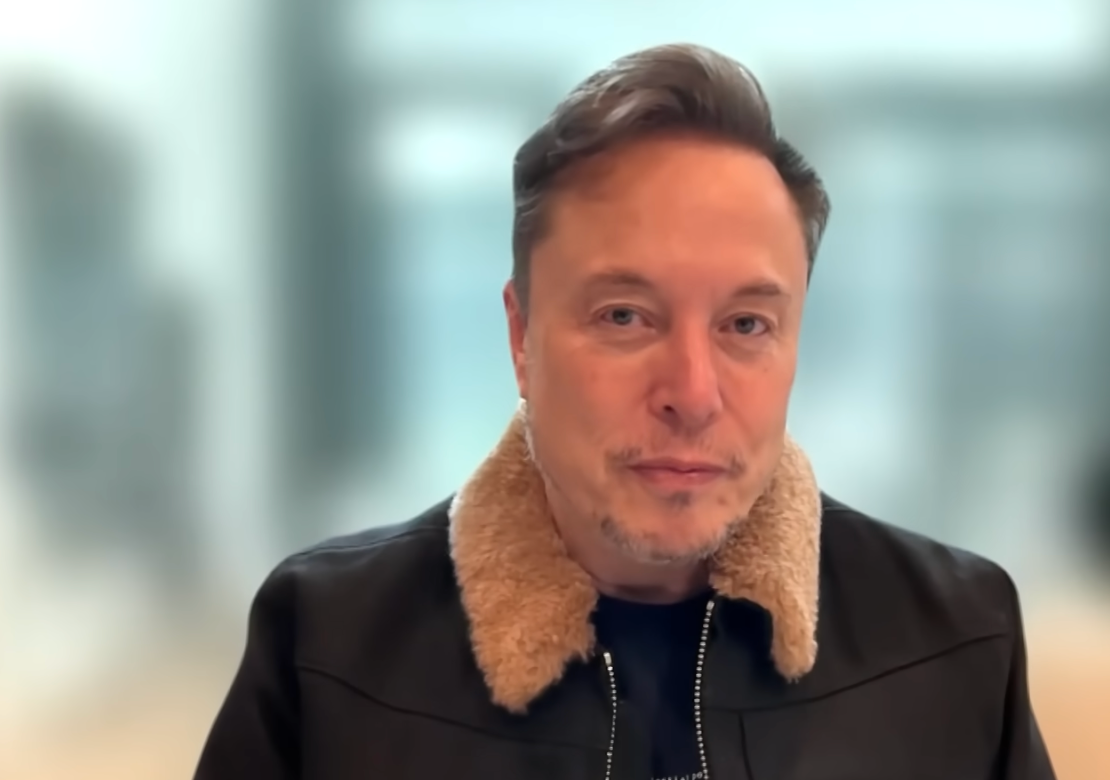 Elon Musk with short hair in a jacket with a fluffy collar, looking directly at the camera. Blurred background