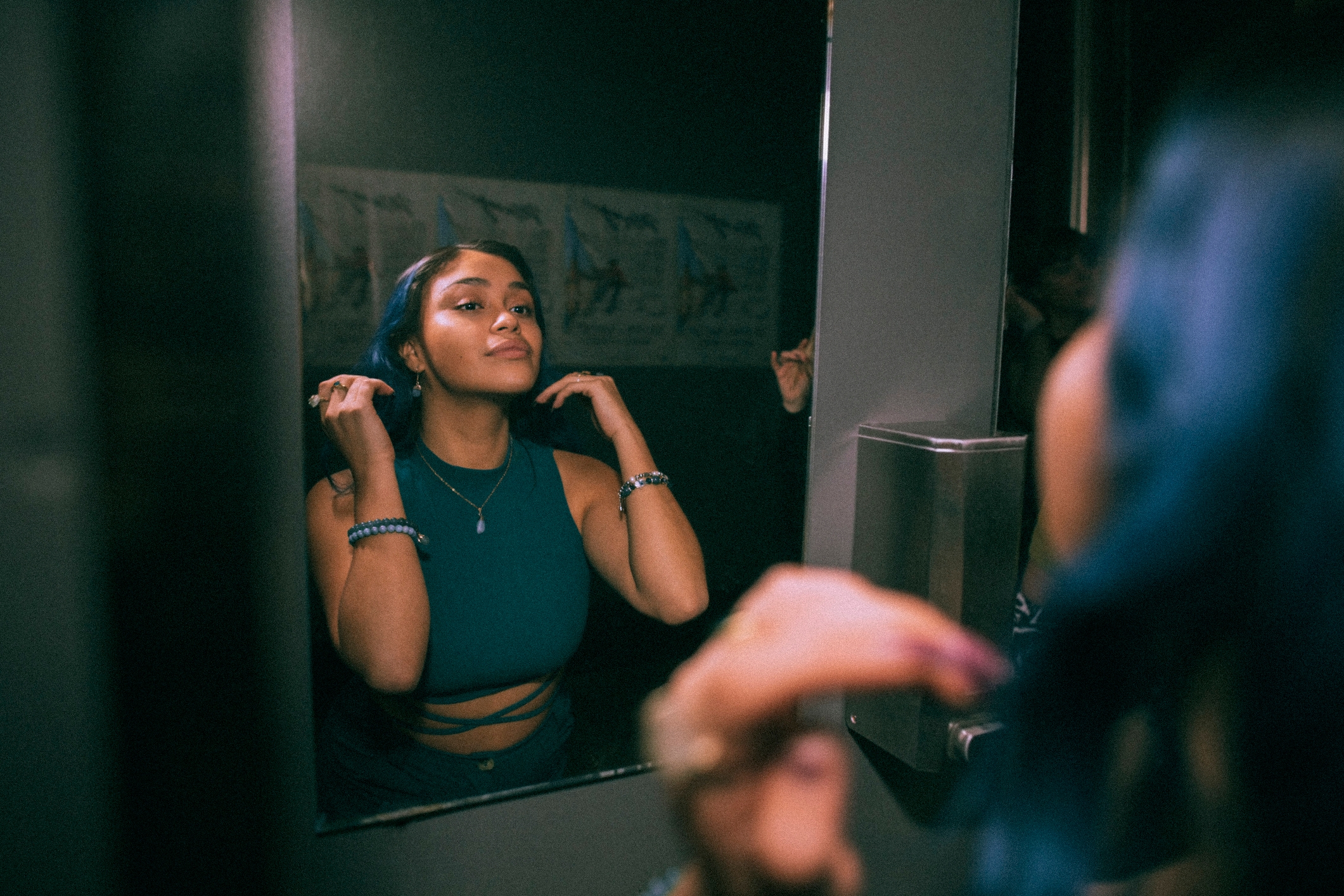Person adjusting hair in front of a mirror, wearing a sleeveless top and bracelets, looking content and confident