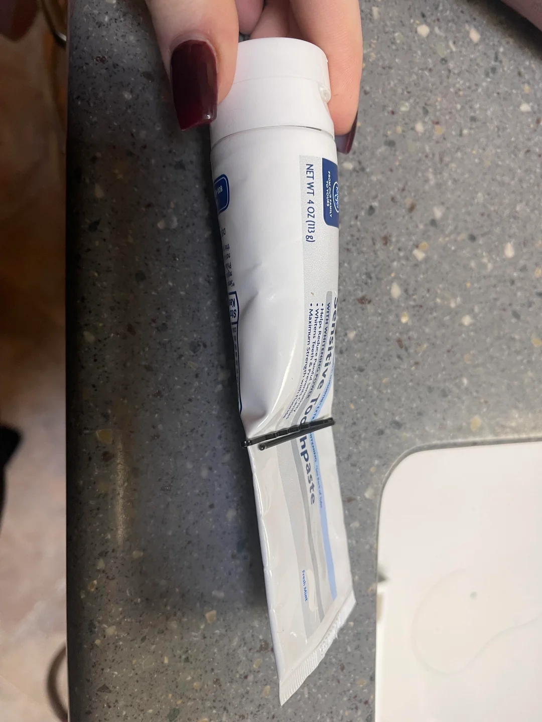 A hand holding a nearly empty toothpaste tube secured with bobby pins to squeeze out the remaining paste