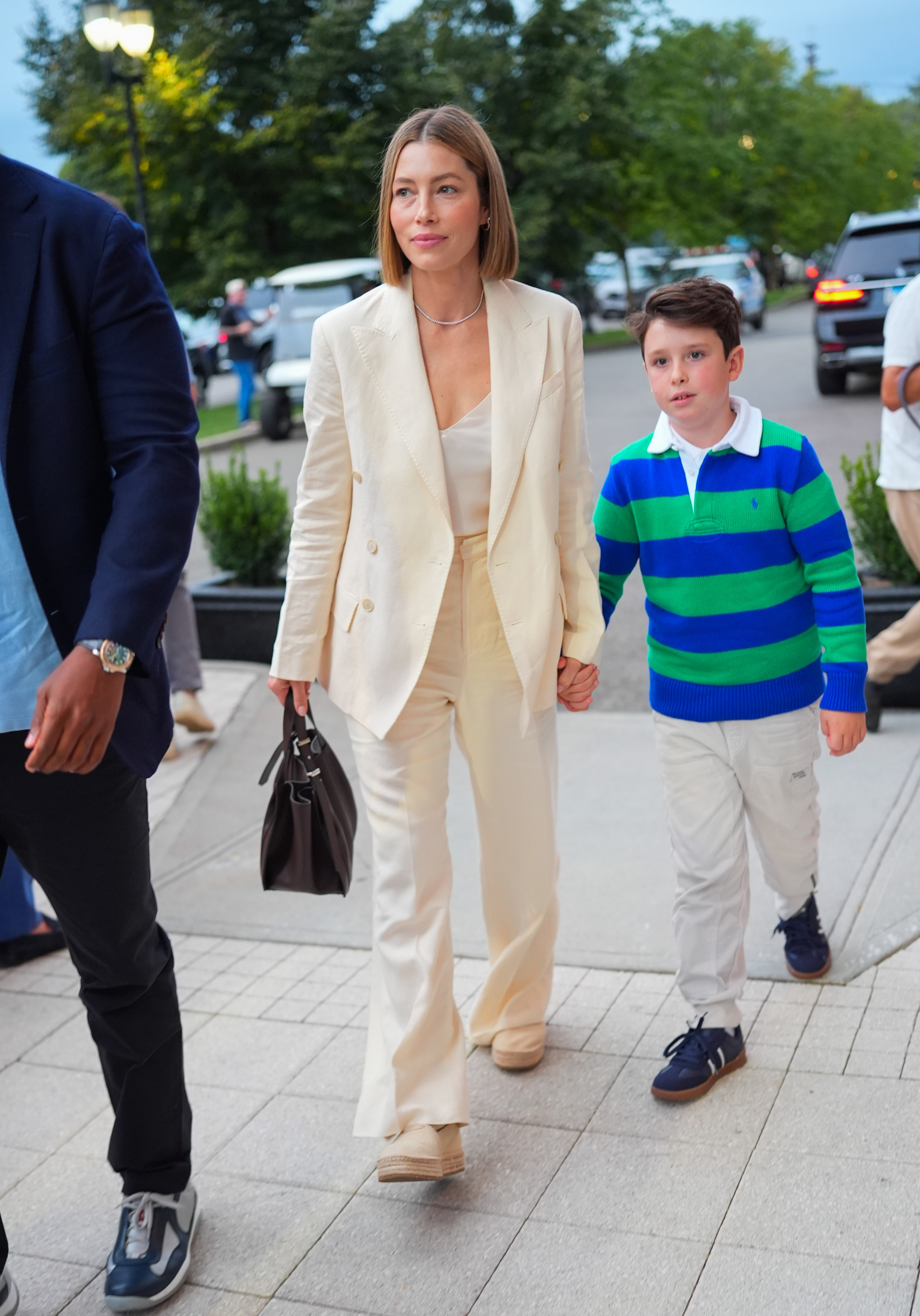 Jessica Biel in a stylish cream suit walks hand-in-hand with a young boy wearing a green-striped sweater. They are outdoors with people and cars in the background
