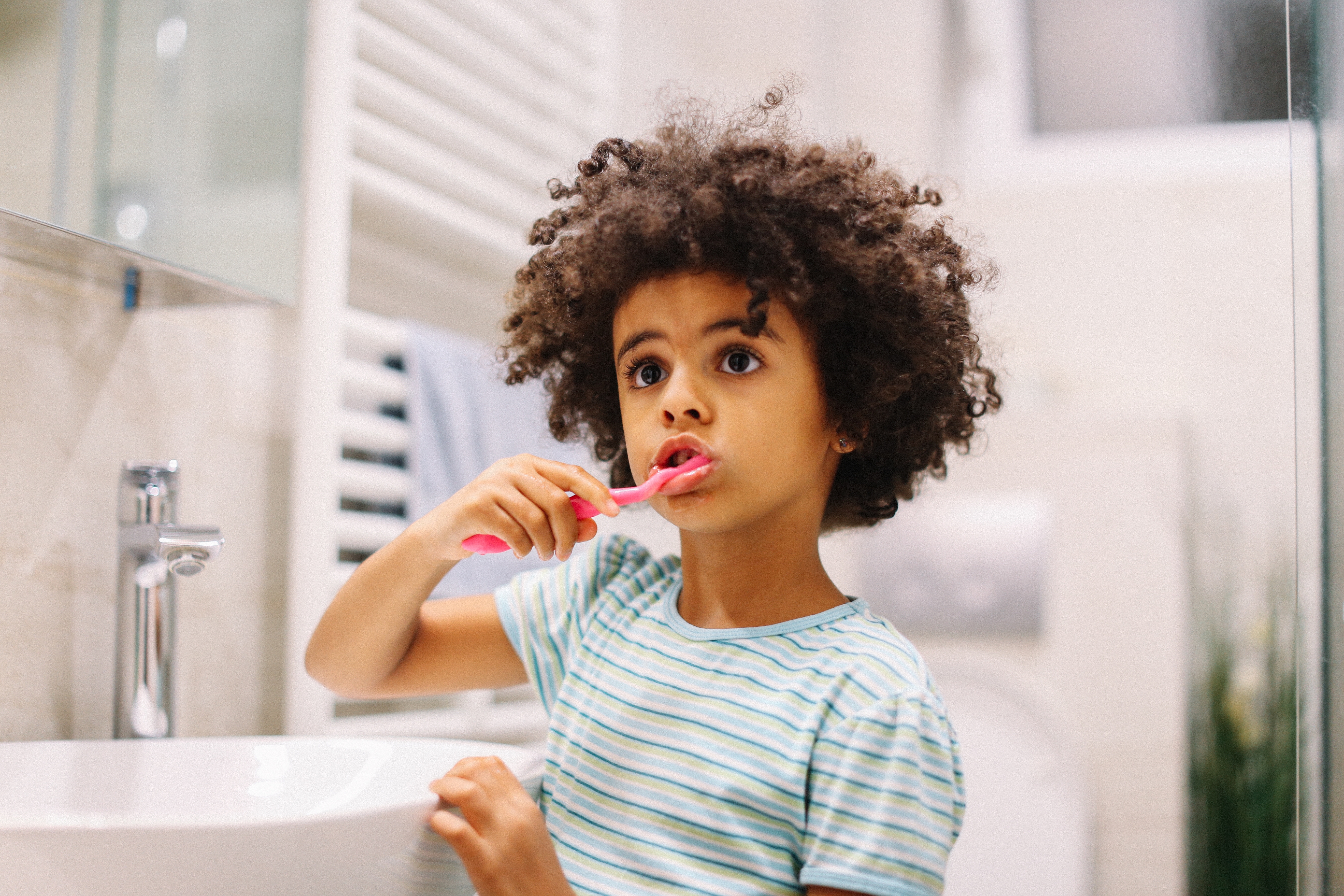 Child in striped shirt brushing teeth in a bathroom, looking thoughtfully into the distance