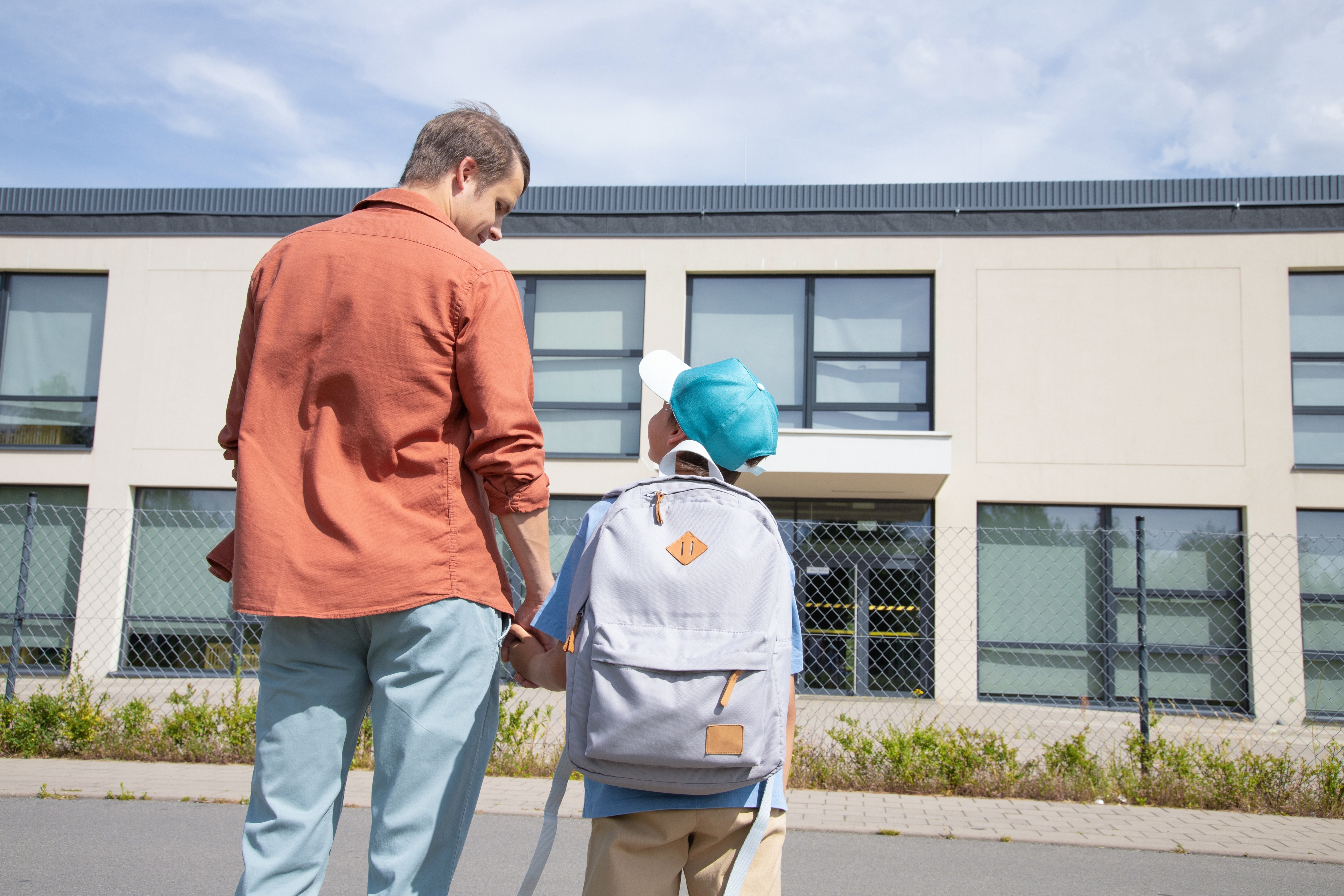 Man and child with backpack holding hands, standing outside a school building