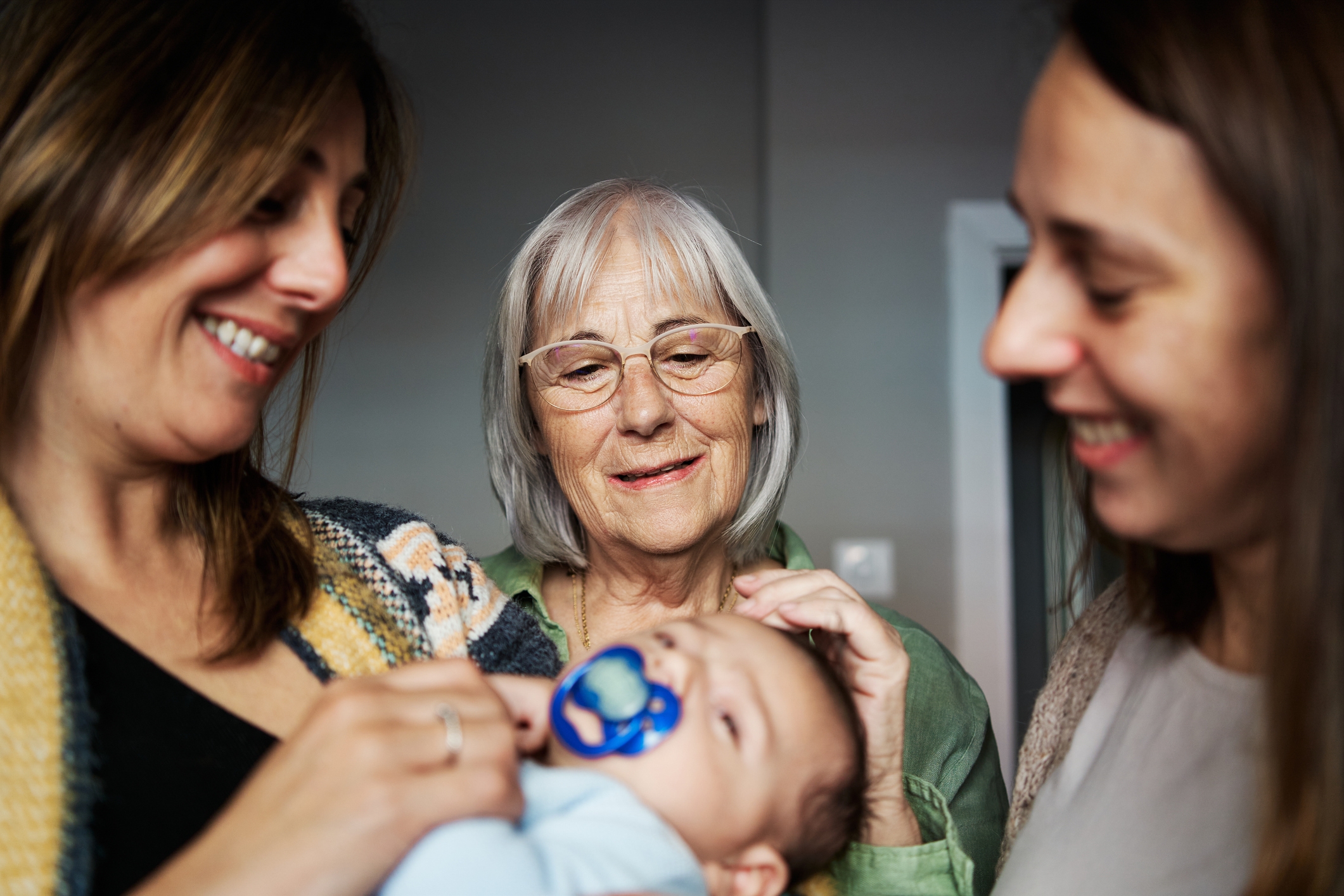 Three women smiling at a baby with a pacifier; the older woman in glasses gently touches the baby's head