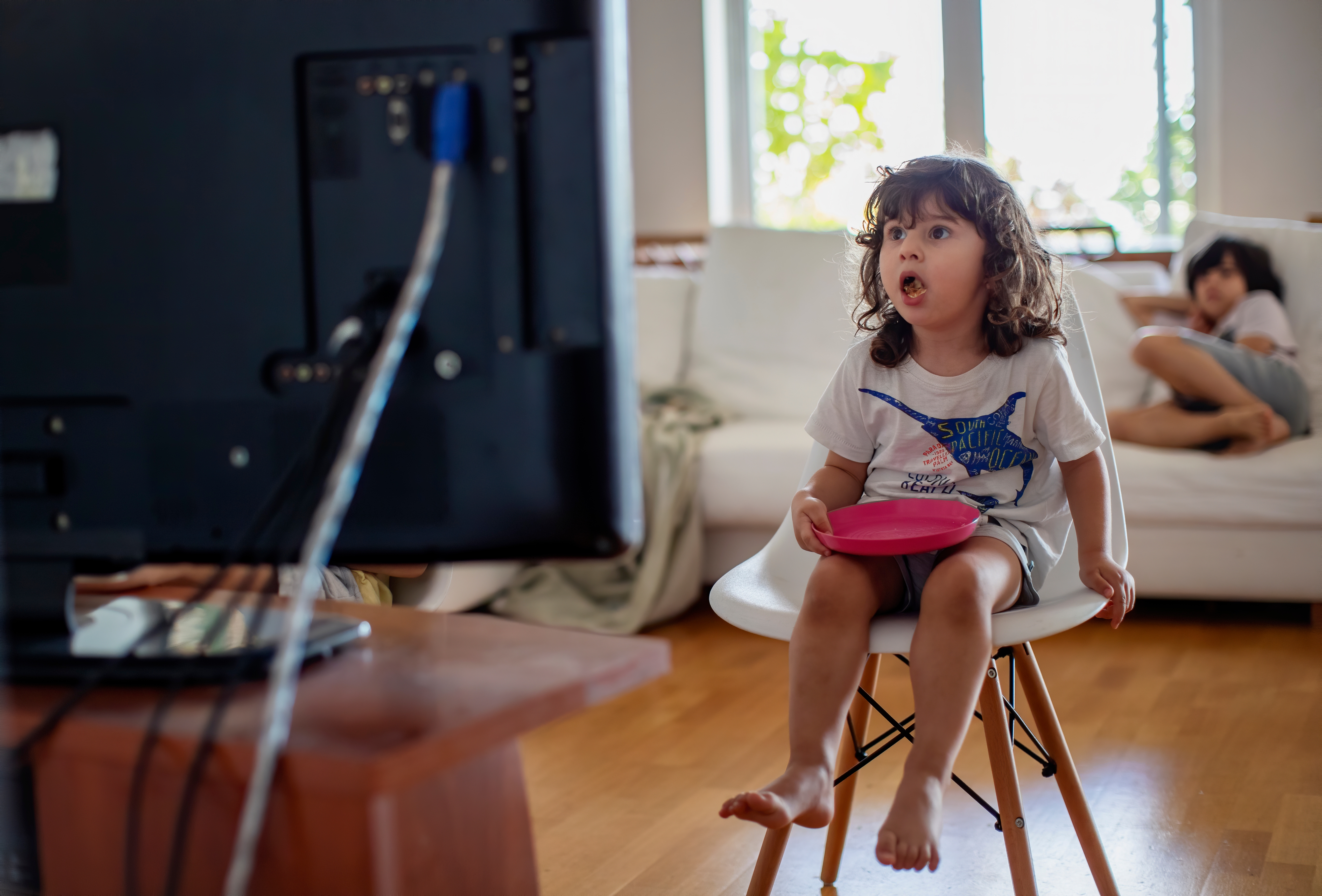 Child sitting on a chair watching TV, holding a bowl, with another child lying on a couch in the background. Living room setting
