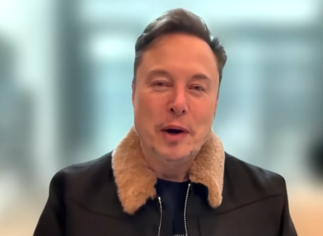 Elon Musk speaking to the camera, wearing a jacket with a fur collar in a blurred indoor setting