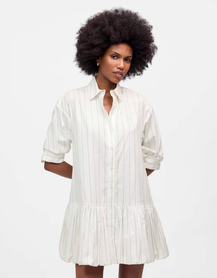 Person wearing a button-down, striped garment formal with rolled sleeves, posing with confidence