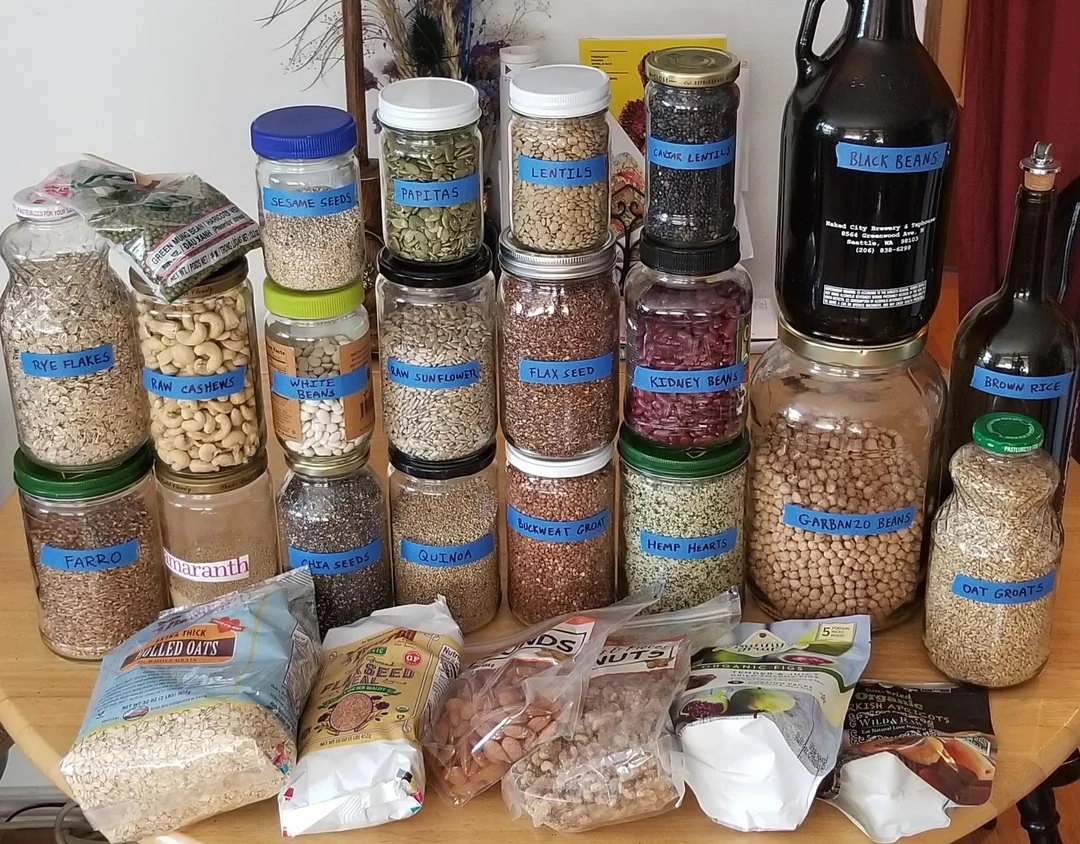 Jars and bottles filled with various grains, nuts, and seeds on a table, showcasing a diverse collection of pantry staples
