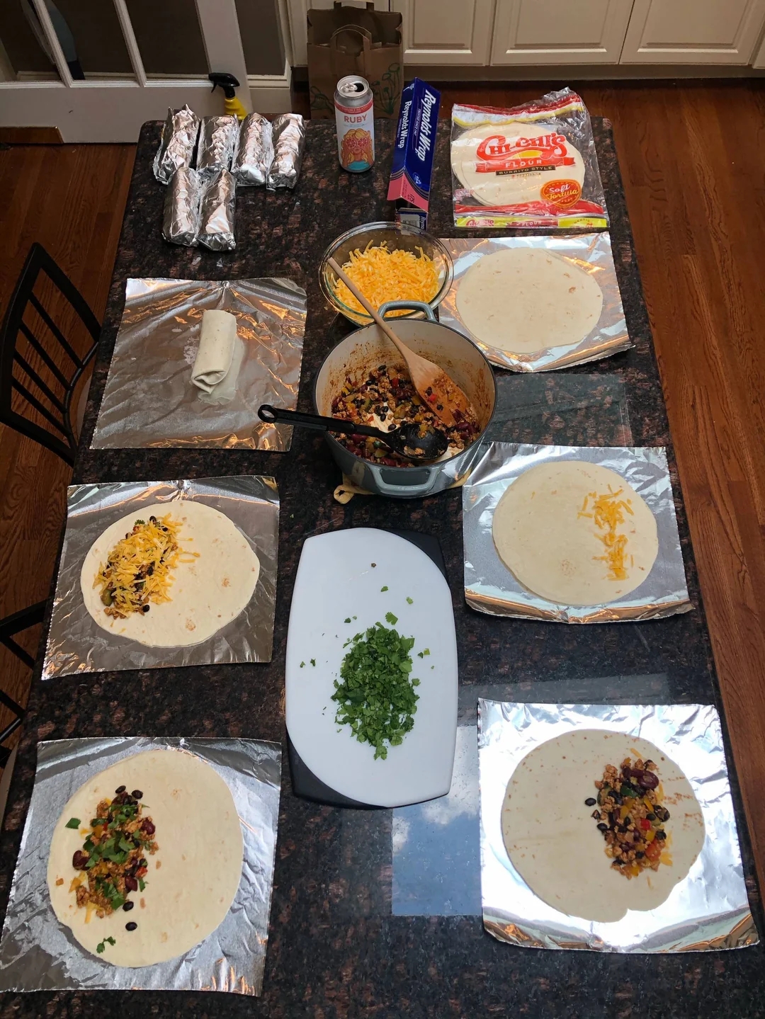 Kitchen counter with tortillas being filled with beans, cheese, and vegetables for burrito preparation. Prepared burritos wrapped in foil are visible