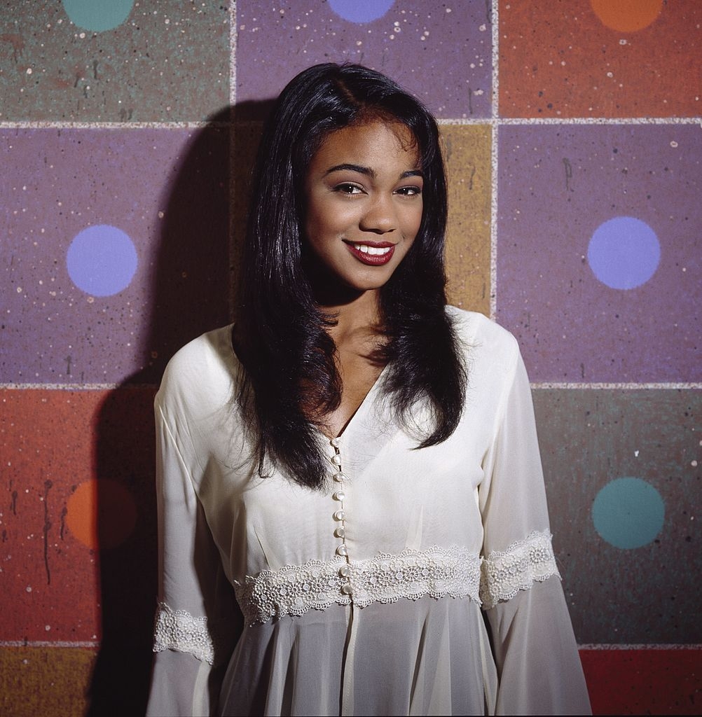 Person posing in front of a textured wall, wearing a buttoned dress with lace details, smiling at the camera