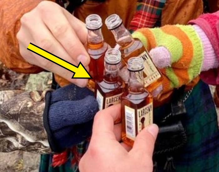 Person in outdoorsy attire, wearing a kilt and hat, clinks mini bottles with others while hiking on a rocky trail