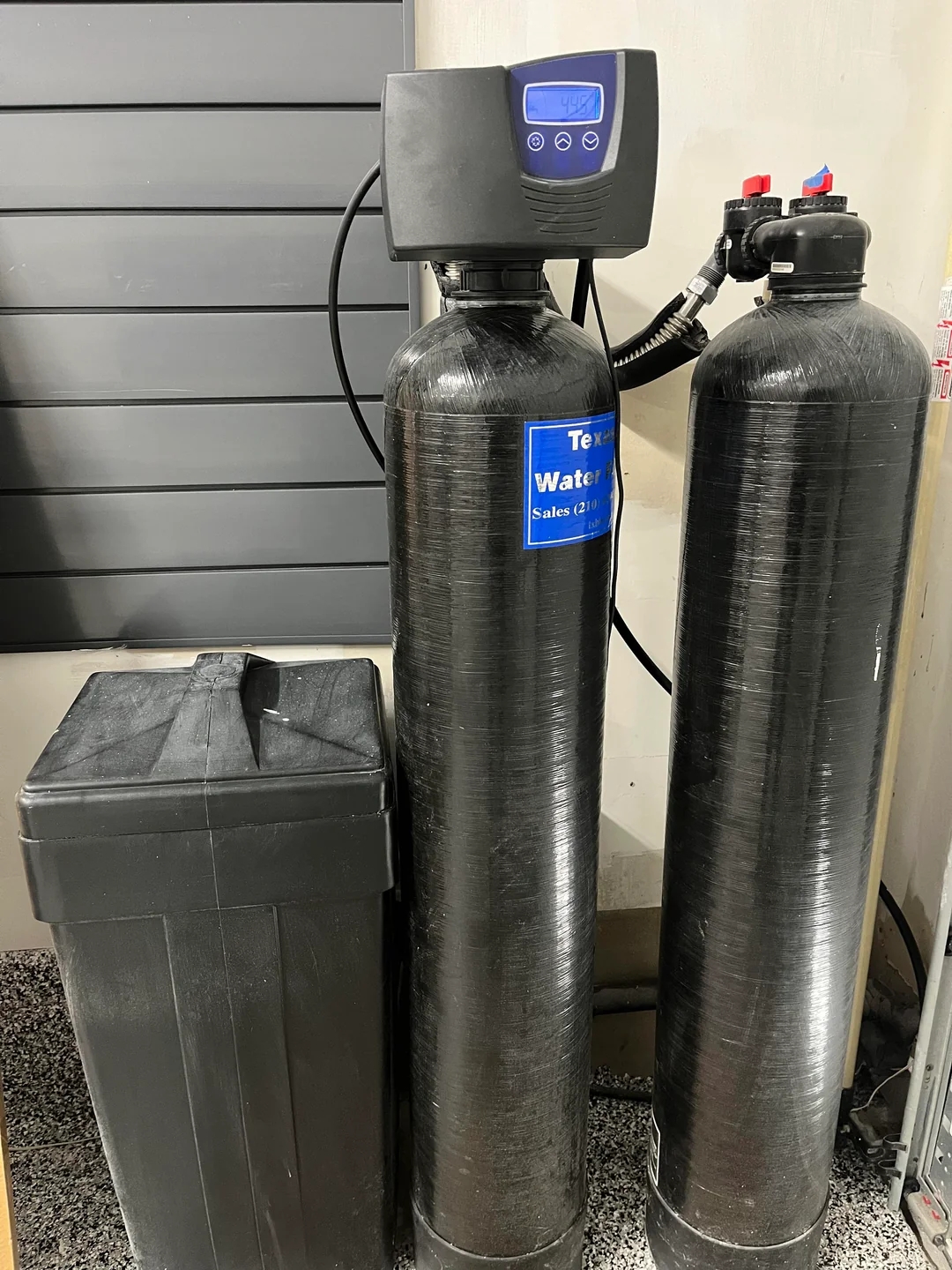 Two black water filtration tanks and a storage bin placed against a wall, used for purifying and softening water in a home or business setting