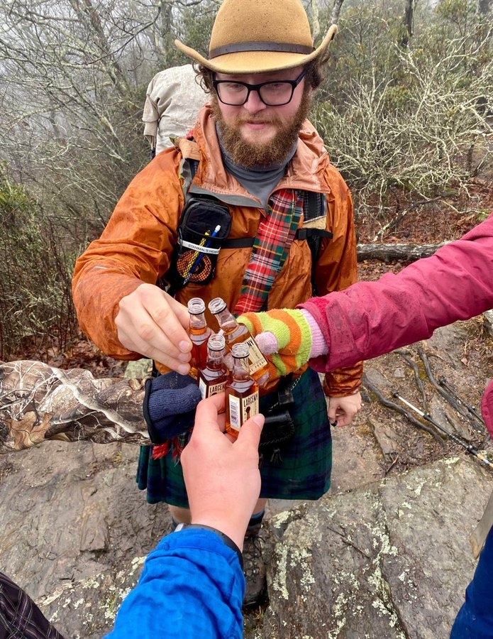 Group of hikers in outdoor attire toasting with mini bottles on a rocky path in a forest