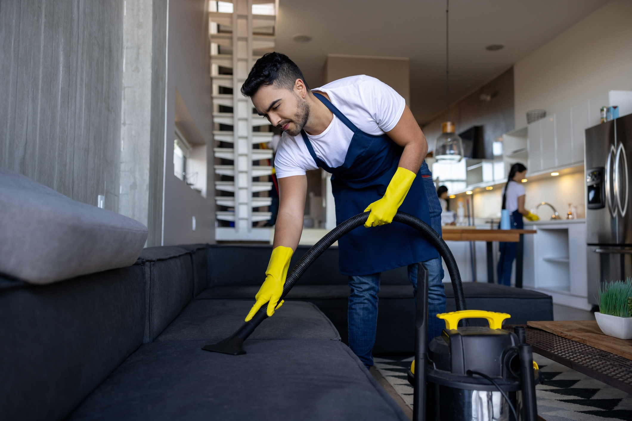 Person in an apron and gloves vacuuming a sofa in a modern home; another person is cleaning in the background
