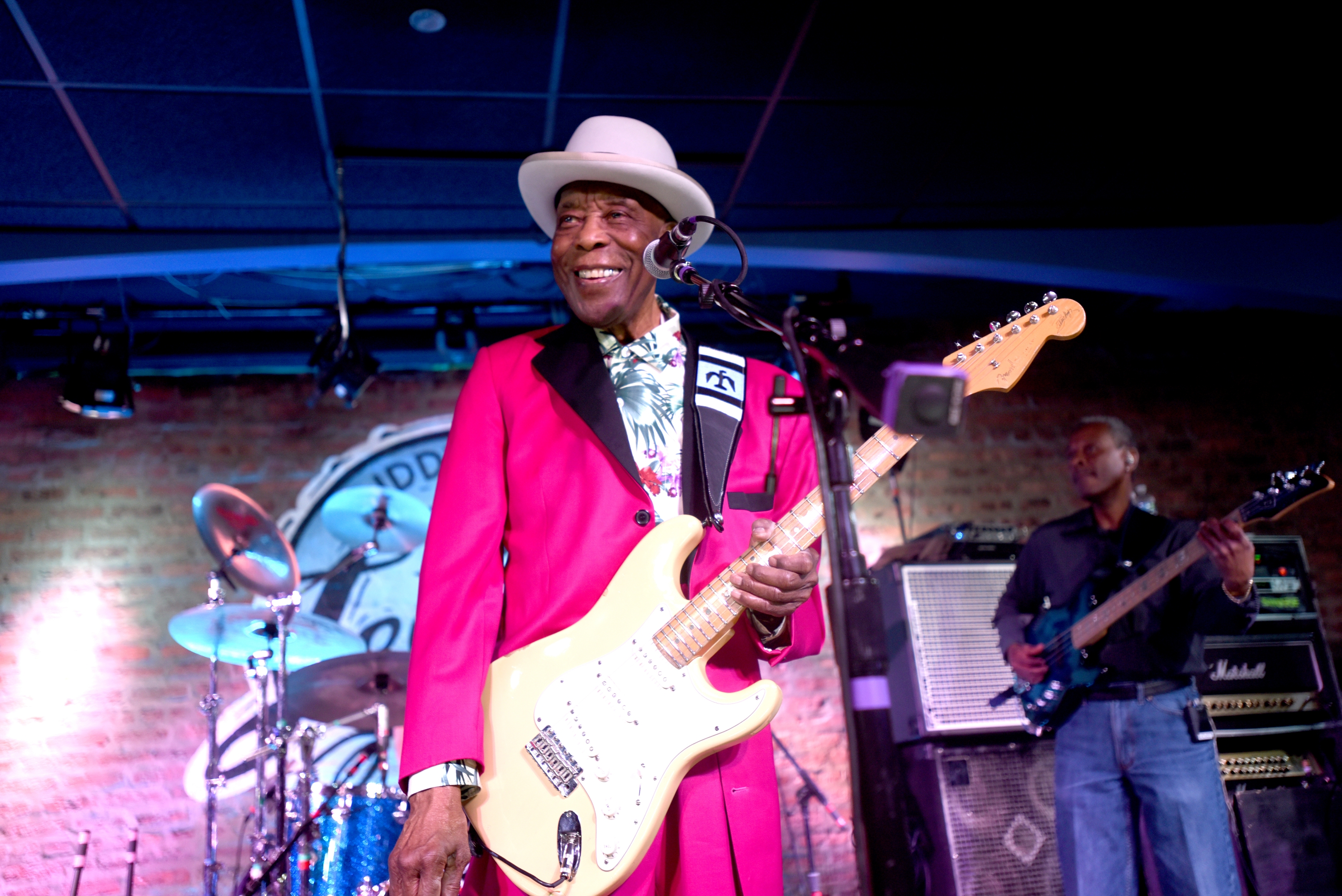 Buddy Guy performing on stage wearing a vibrant suit and hat, playing an electric guitar, with a band in the background