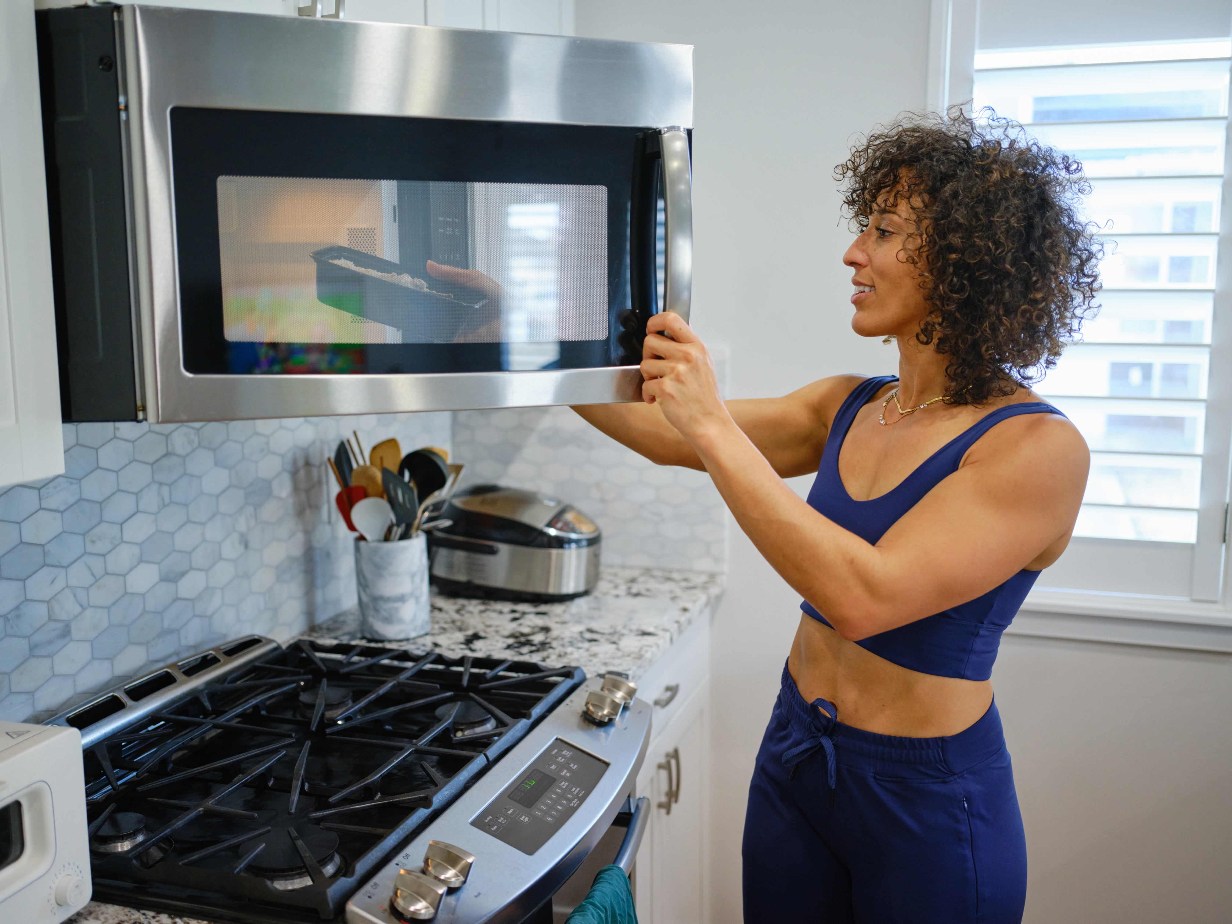 Person with curly hair in workout attire opens a microwave above a stove in a modern kitchen