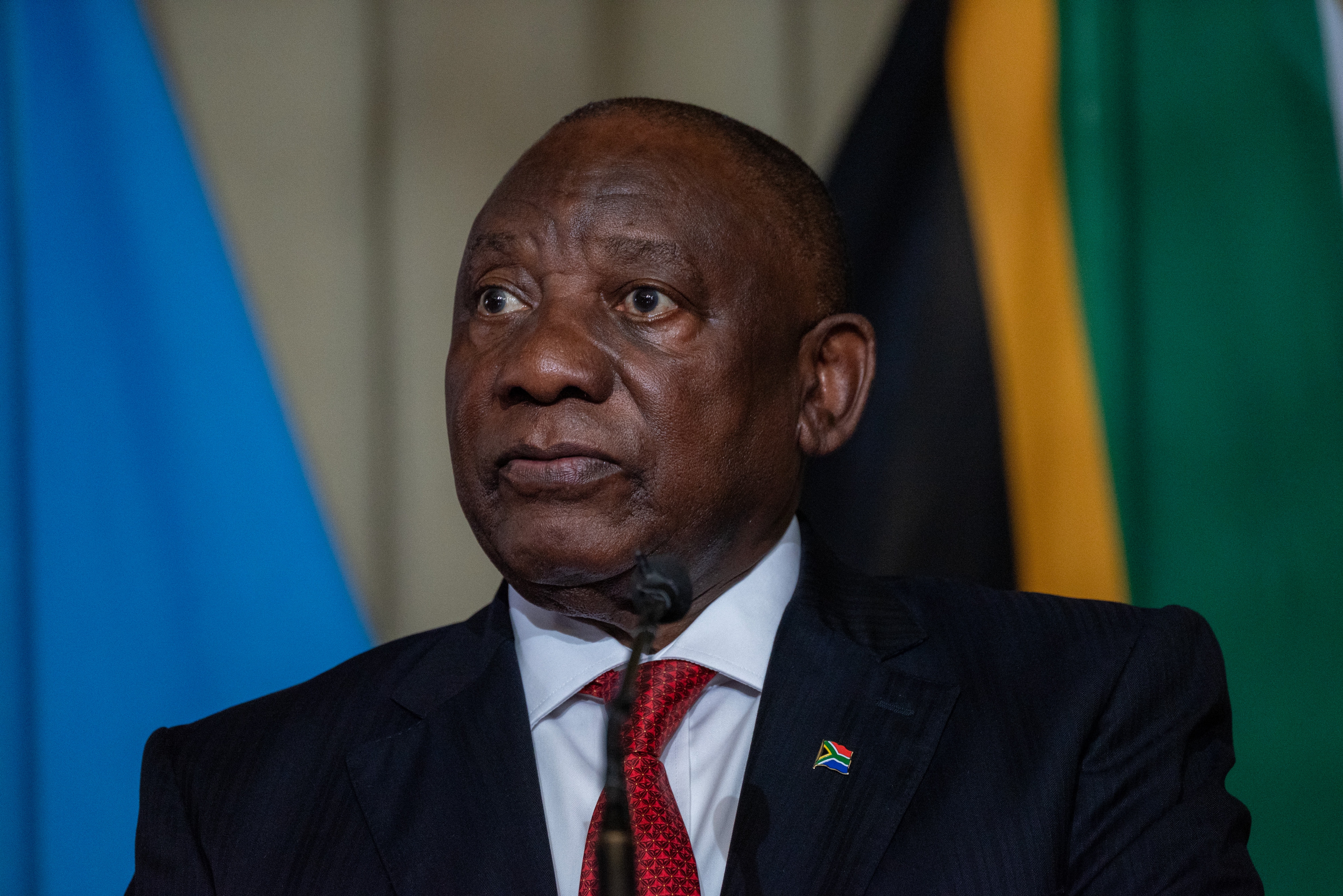 Cyril Ramaphosa in a suit with a South Africa flag pin speaks at a podium, with flags in the background