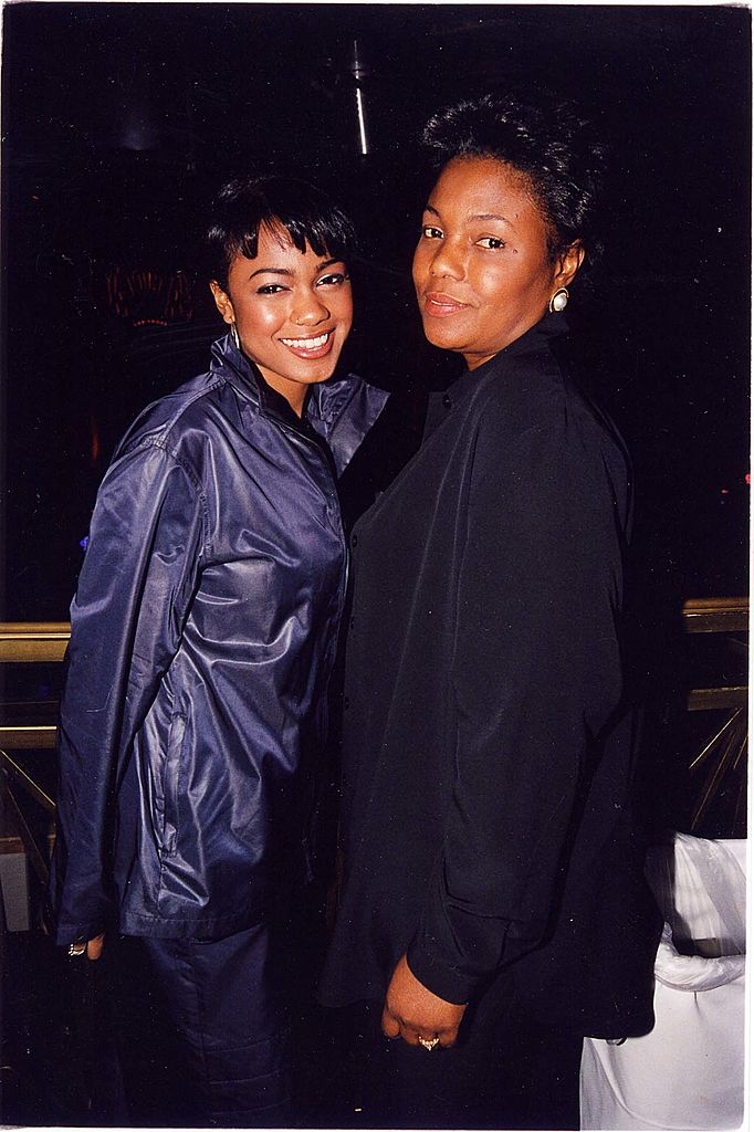 Two women smiling at an event, one in a shiny jacket, the other in a matte outfit. They are posing together in a casual setting