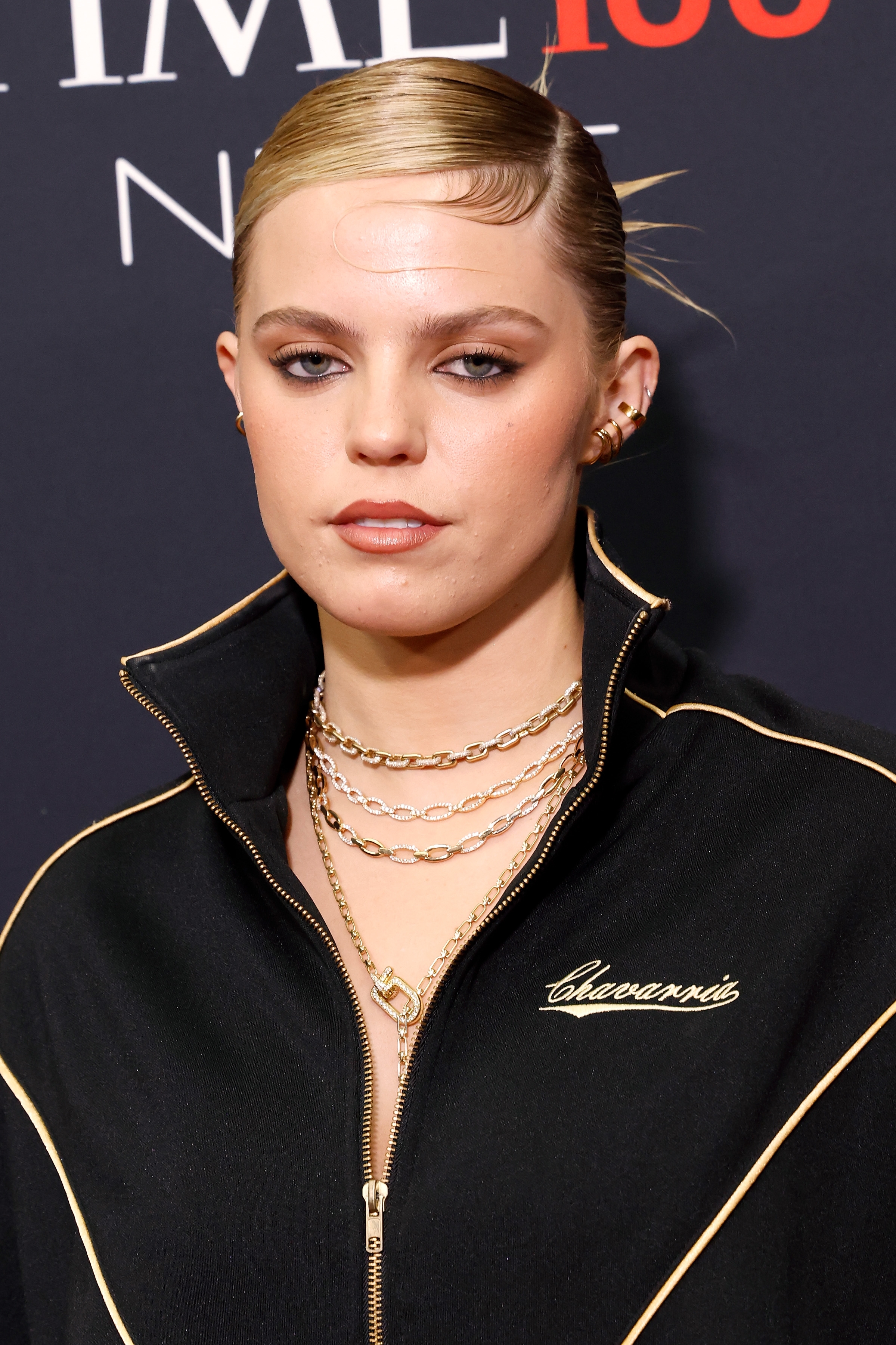 Reneé Rapp in a track jacket with layered gold necklaces at an event. Hair is styled back with a sleek appearance