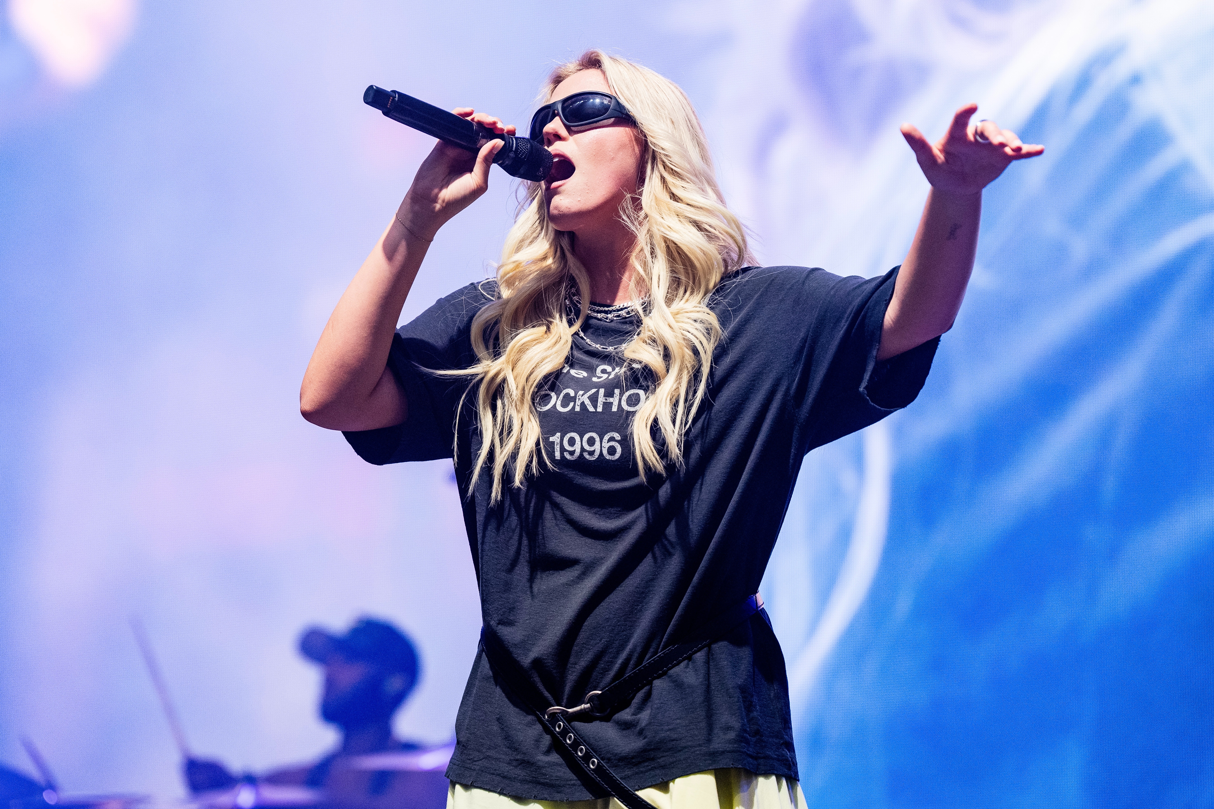 Renee Rapp wearing oversized shirt and sunglasses performs energetically on stage, holding a microphone with a band playing in the background