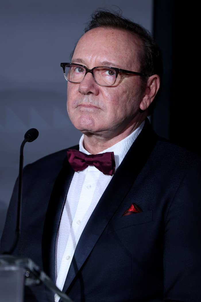 Kevin Spacey person in a suit and bow tie stands at a podium, looking upwards