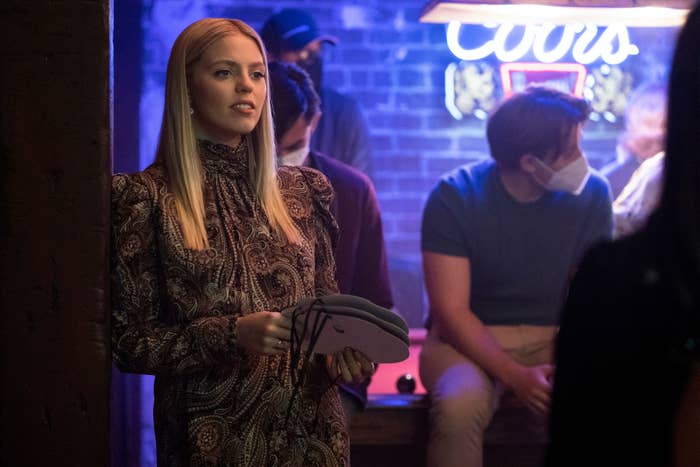 Renee Rapp in patterned, long-sleeved clothing holds horseshoes in a dimly lit bar with people seated in the background