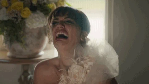 A person stands indoors laughing, wearing a bridal dress with floral details