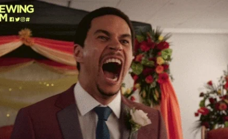 Person in a suit with a boutonniere yelling in excitement at a wedding ceremony, with floral decorations in the background