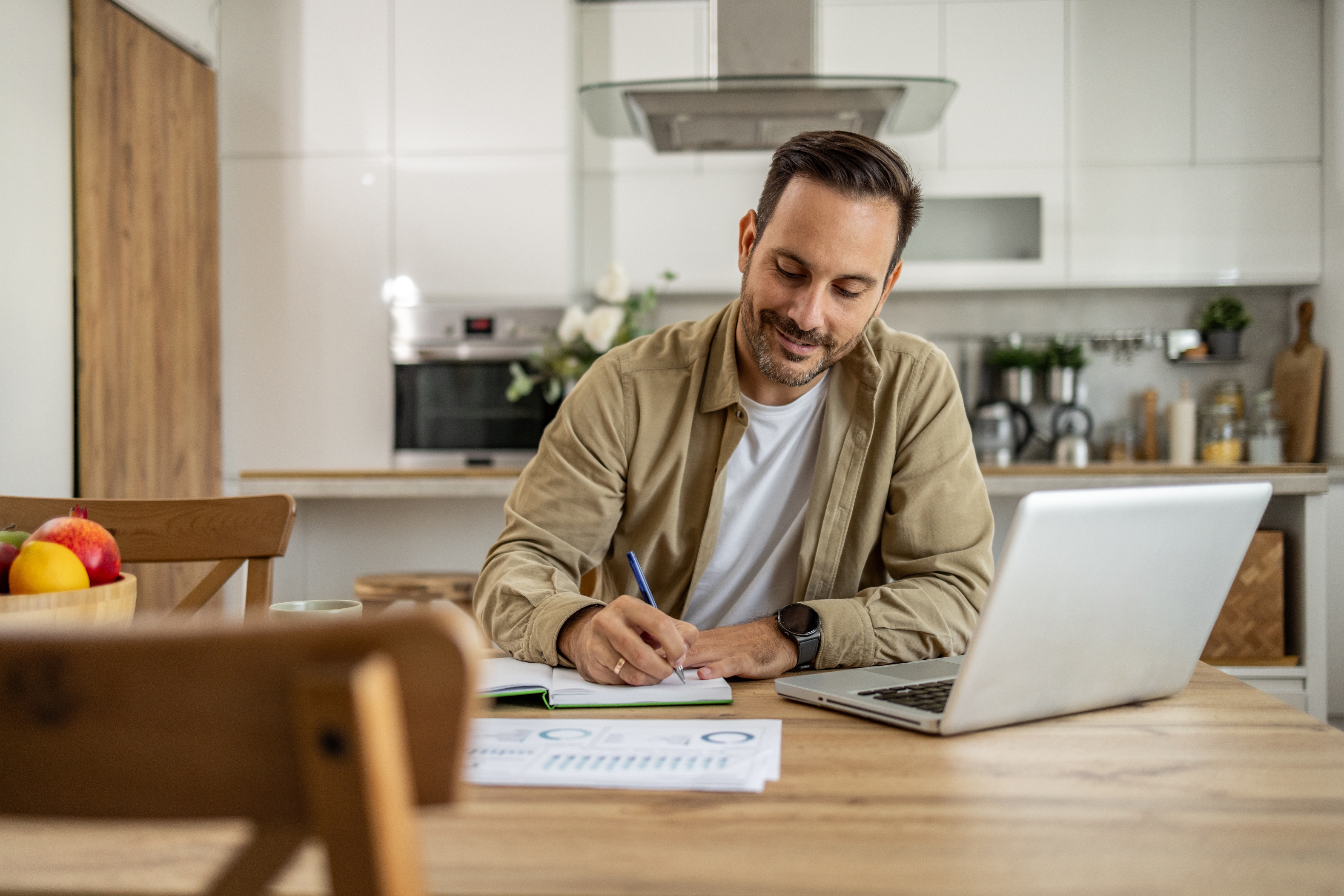 Man smiling, writing in notebook at kitchen table with laptop, papers, and mug
