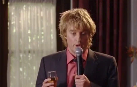 Person in a suit holding a microphone and a glass, speaking at a wedding event in an indoor setting