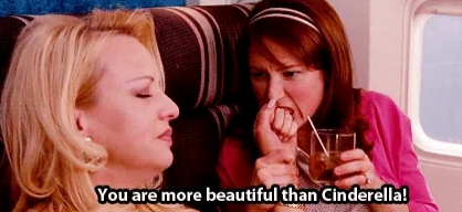 Two women seated on an airplane; one holds a drink. Subtitled text reads, "You are more beautiful than Cinderella!"