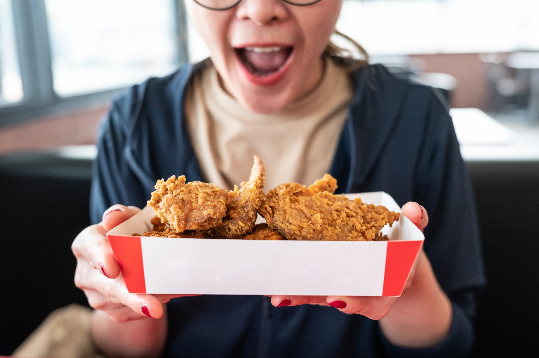 Person excitedly holding a box of fried chicken, about to eat, inside a casual dining setting