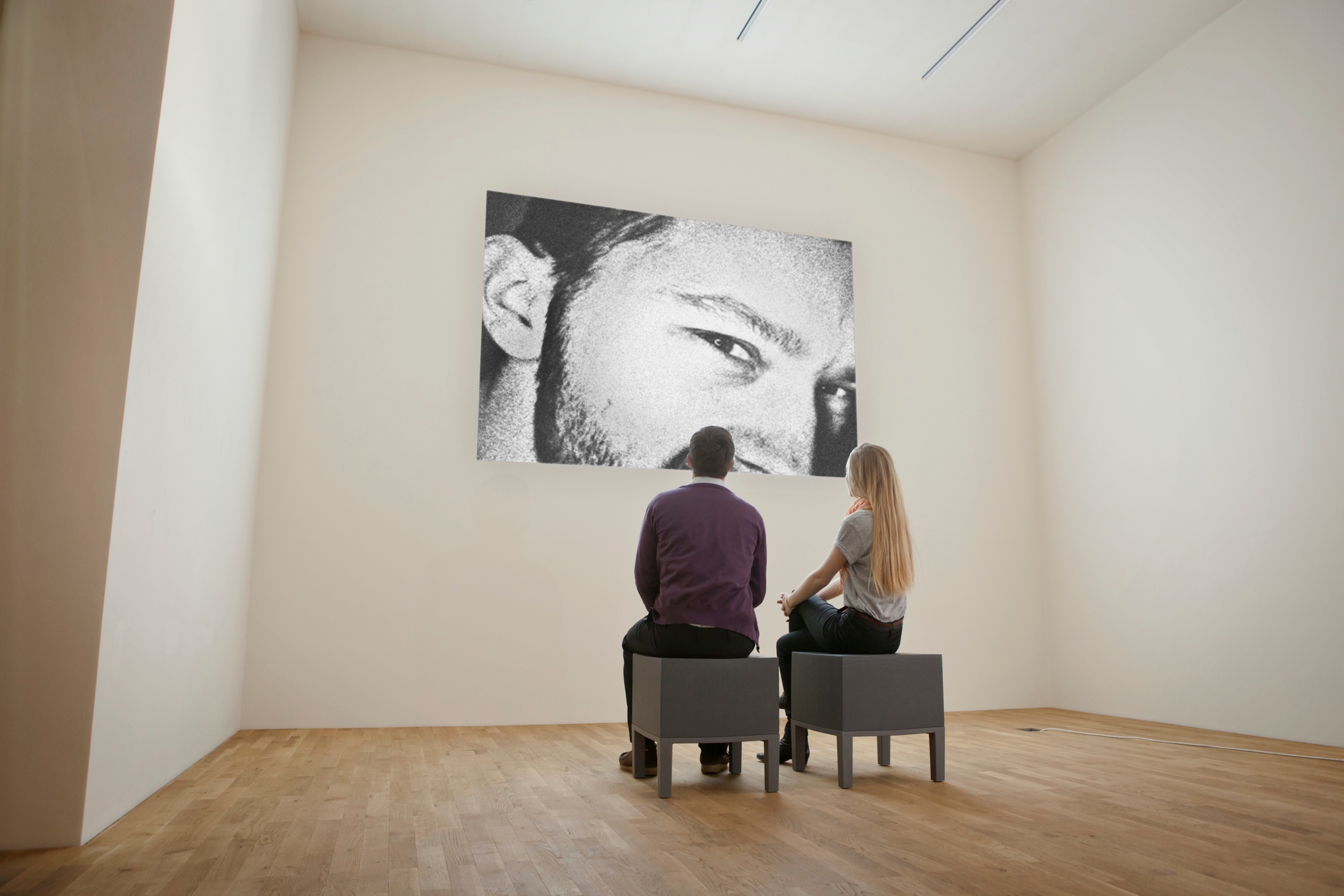Two people sit on benches, observing a large black-and-white portrait of a man's face in a minimalist gallery setting