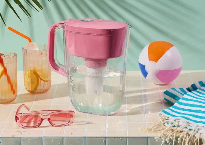 A Brita water pitcher with pink lid is on a table near two drinks, sunglasses, a beach ball, and a towel. Text: "Summer hydration made easy with Brita."