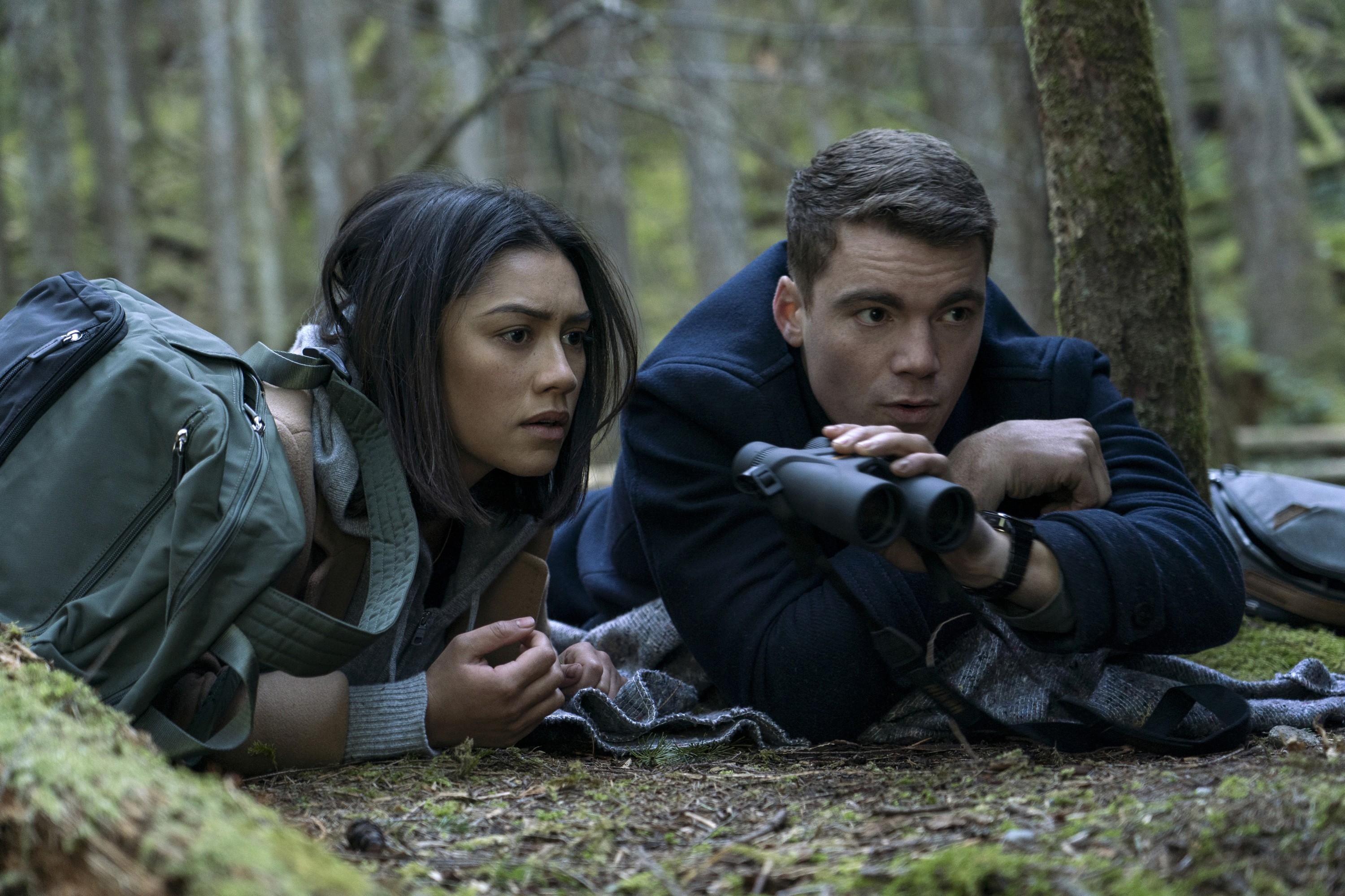 Two people lie on forest ground, one holding binoculars. They look focused and alert, surrounded by trees and greenery