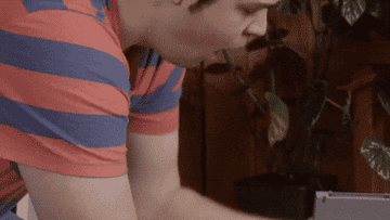 Person in striped shirt types frantically on a laptop, appearing focused and surprised. Plant is visible in the background