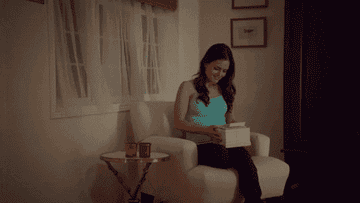 A woman sitting on a chair looks pleasantly surprised while opening a gift box