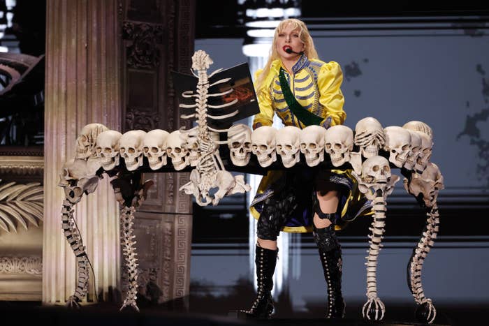 Lady Gaga performs on stage behind a stand decorated with skull motifs, holding a microphone