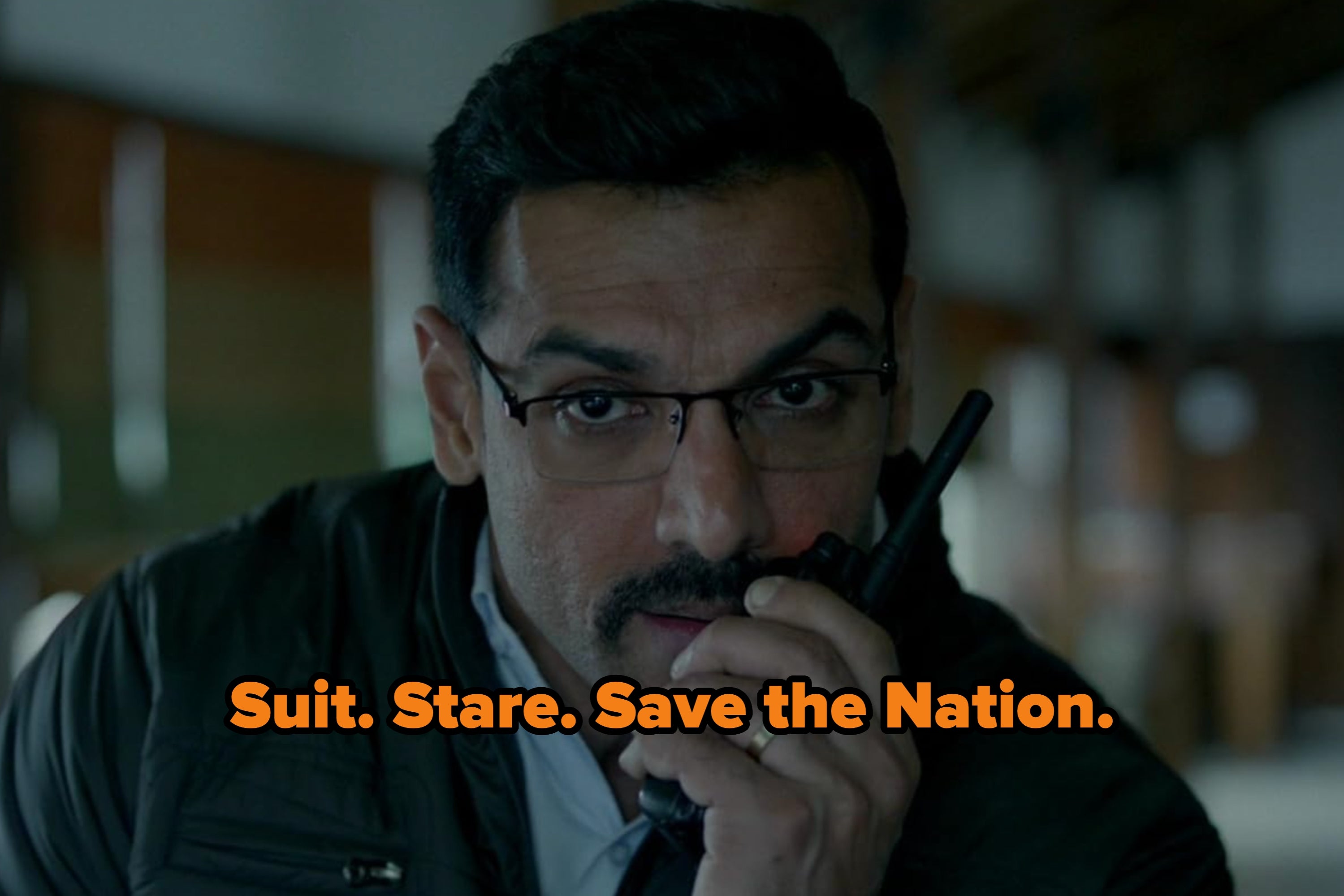 A person with glasses and a mustache is holding a walkie-talkie close to their mouth indoors