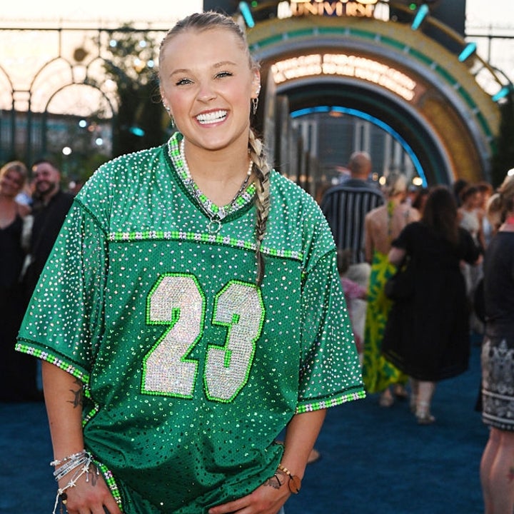 Person in casual football jersey and cargo pants on a red carpet at an outdoor event with an entrance sign in the background